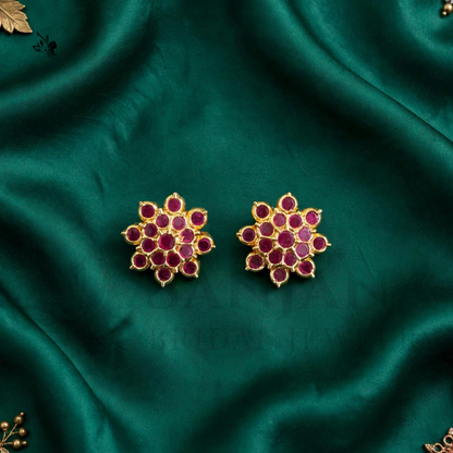 A pair of gold-toned floral stud earrings with deep red stones, displayed on a draped dark green satin fabric.