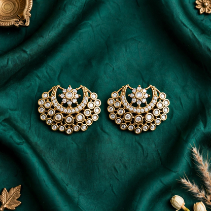 A pair of ornate gold crescent-shaped earrings with intricate white stone detailing, displayed on a draped dark green fabric background with decorative dried grass and gold-toned props.