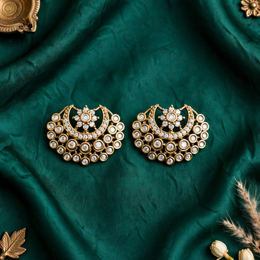 A pair of ornate gold crescent-shaped earrings with intricate white stone detailing, displayed on a draped dark green fabric background with decorative dried grass and gold-toned props.