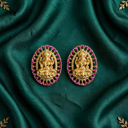 A pair of oval gold-toned jewelry pieces featuring a central deity motif surrounded by pink gemstones, displayed on a draped dark green satin fabric.