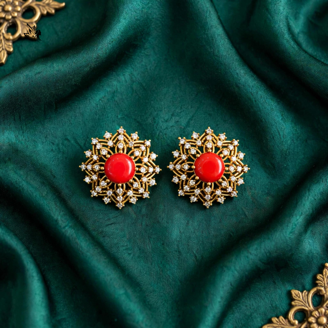 A pair of ornate gold-toned earrings with round red centers and small clear stones, displayed on draped dark green fabric.