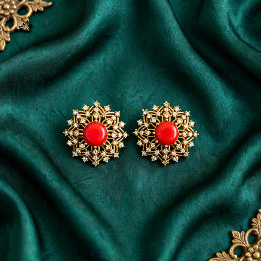 A pair of ornate gold-toned earrings with round red centers and small clear stones, displayed on draped dark green fabric.