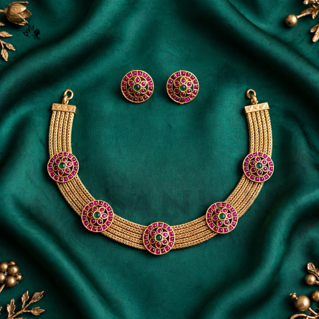 Gold-toned traditional necklace set with matching stud earrings, decorated with pink and green stone circular motifs, arranged on draped emerald green fabric with small gold decorative accents.