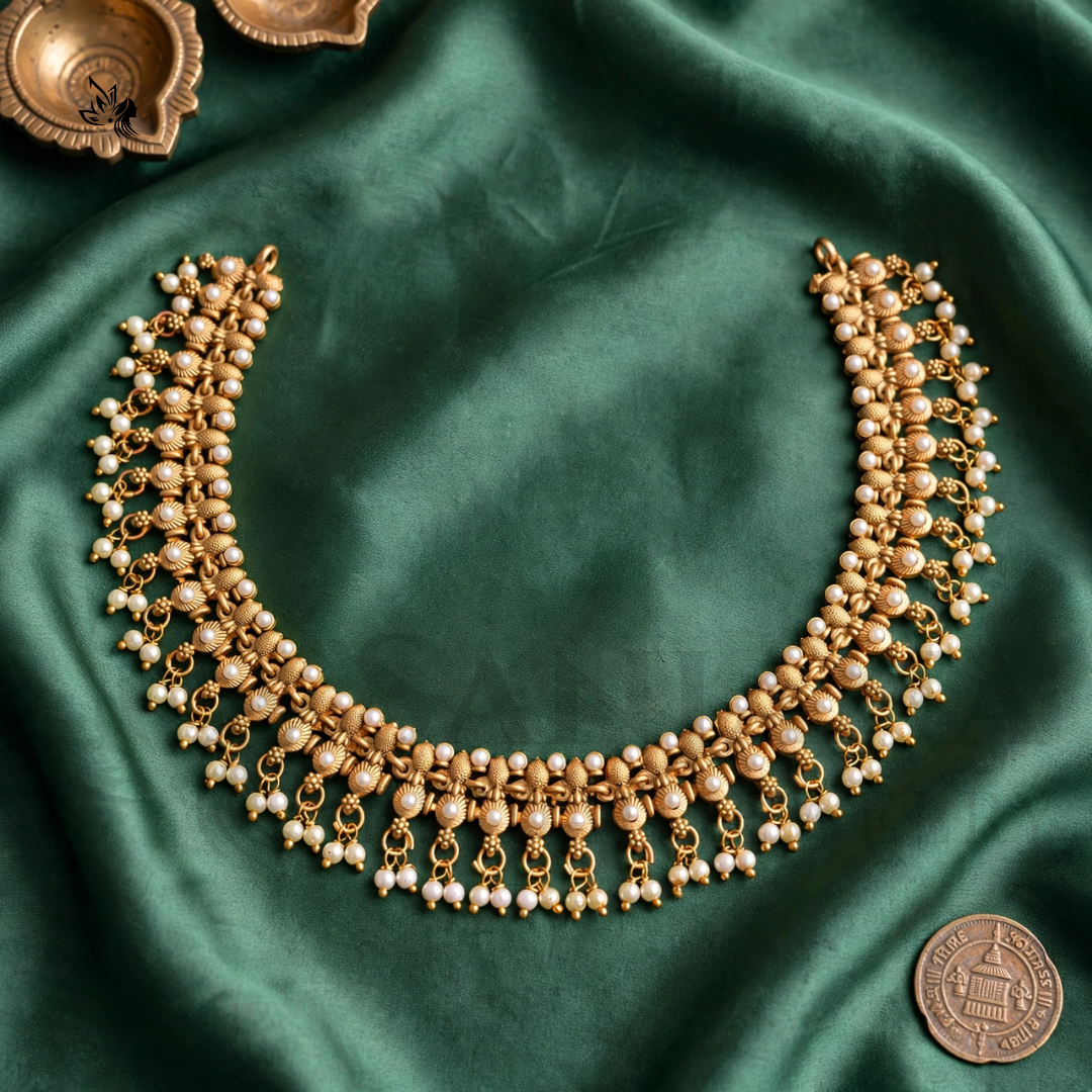An ornate gold-toned necklace with rows of small white pearl-like beads and dangling bead chains, arranged on draped green satin fabric. A coin is placed near the lower right for scale, and decorative brass objects appear at the top corners.