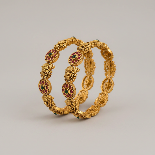 A pair of ornate gold-plated bangles with intricate filigree patterns and red and green enamel floral details, displayed on a neutral gray background.