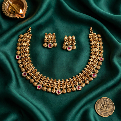 An ornate gold-toned traditional necklace set with matching earrings, displayed on draped emerald green satin fabric. The jewelry features intricate beadwork and small pink gemstone accents.