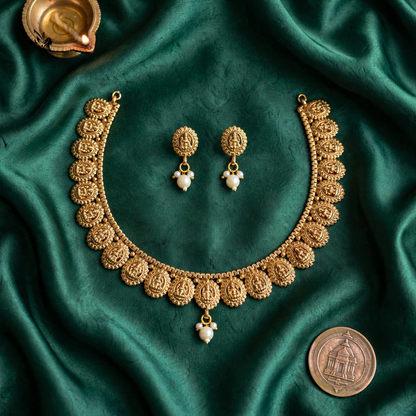 An ornate gold-toned jewelry set arranged on draped emerald green satin fabric, including a necklace, matching earrings, and a coin placed nearby for scale.