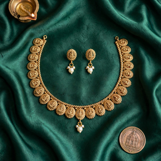 An ornate gold-toned jewelry set arranged on draped emerald green satin fabric, including a necklace, matching earrings, and a coin placed nearby for scale.