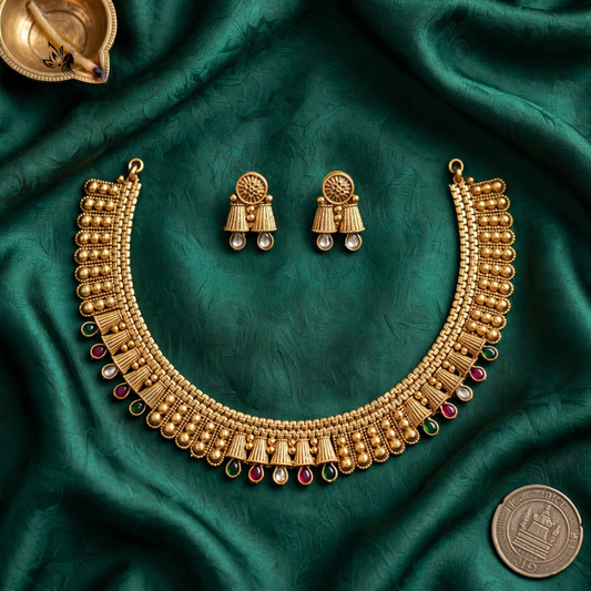 Gold-toned traditional Indian jewelry set featuring a wide beaded necklace and matching earrings, arranged on draped emerald green fabric with a coin and brass oil lamp as props.
