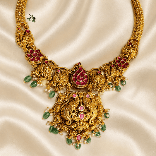 An ornate gold-toned traditional necklace with red and pink stone embellishments and green teardrop accents, displayed on a cream satin fabric background.