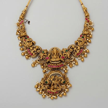 An ornate gold-toned traditional necklace with intricate filigree, red enamel accents, green stone details, and small pearl-like beads, displayed on draped cream satin fabric.