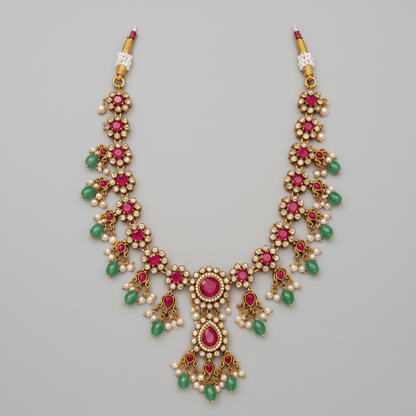 A gold-plated traditional Indian Kundan necklace with vibrant pink gemstones, green stones, and white faux pearls, displayed on a cream satin fabric background with white roses in the corner.
