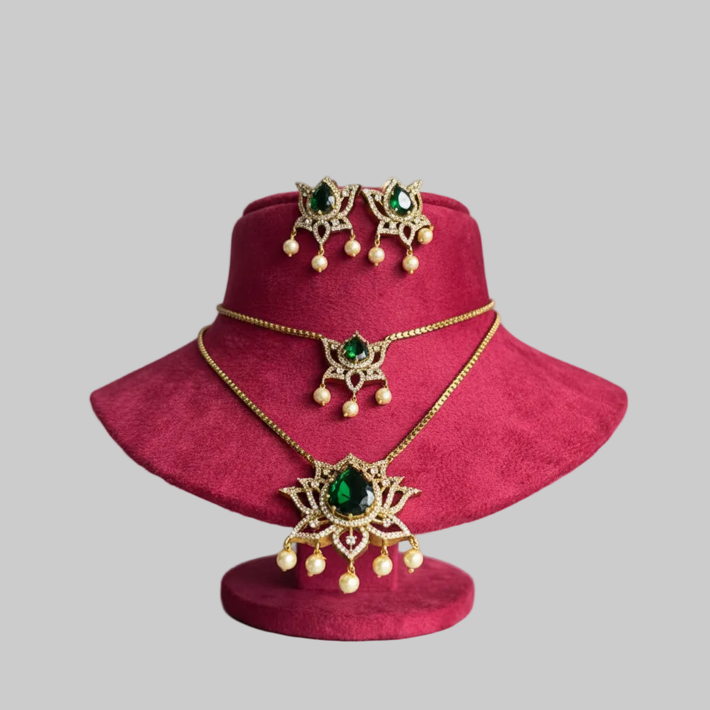 Gold-toned jewelry set with a necklace, matching earrings, and a small pendant, each featuring green gemstone centers and pearl-like drops, displayed on a magenta velvet stand against a light gray background.