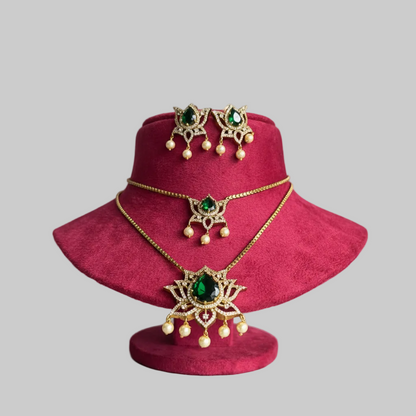 Gold-toned jewelry set with a necklace, matching earrings, and a small pendant, each featuring green gemstone centers and pearl-like drops, displayed on a magenta velvet stand against a light gray background.