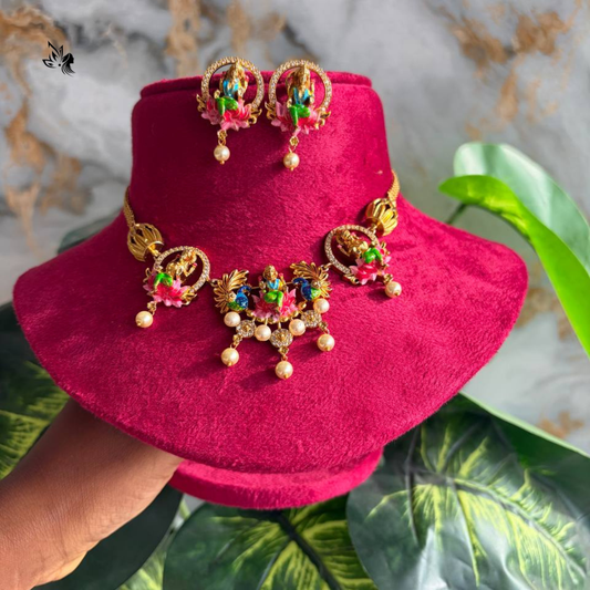 Gold-toned traditional Indian jewelry set including a necklace, earrings, and a matching headpiece, displayed on a bright pink velvet stand. The jewelry features ornate gold filigree, green and red stone accents, and small pearl-like beads.