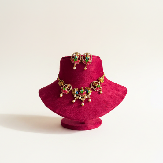 Gold-toned traditional Indian jewelry set featuring a necklace and matching earrings with red, green, and blue stones, displayed on a magenta velvet jewelry bust against a light beige background.
