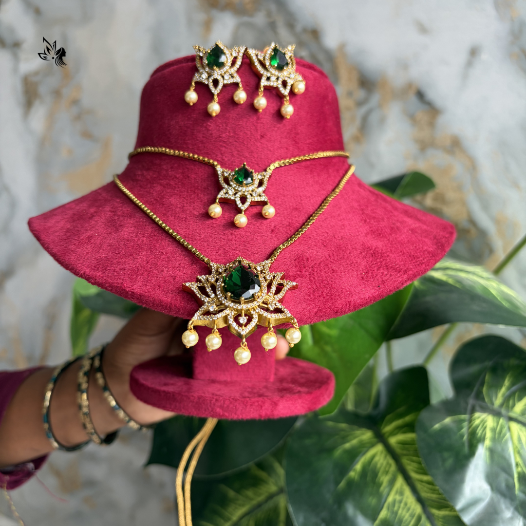 Gold-toned jewelry set with a necklace, earrings, and bracelet displayed on a bright pink velvet hat stand, held by a hand against a marble and green plant background.