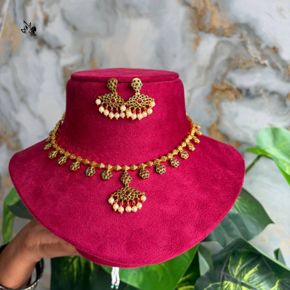 Gold and cream bead jewelry set displayed on a magenta velvet stand, featuring a necklace, matching earrings, and a small pendant, with a hand holding the stand against a marble and green plant background.