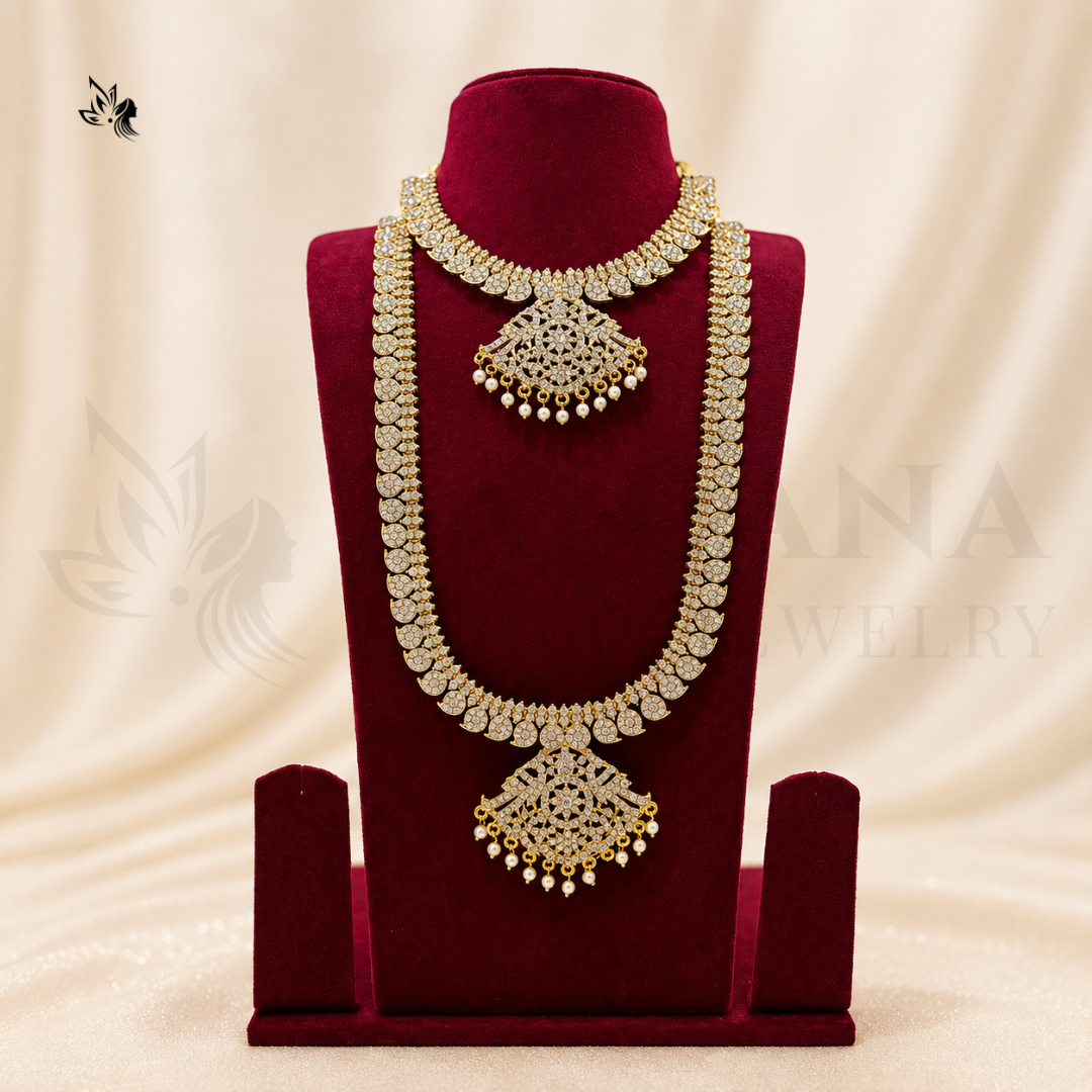An ornate gold-toned traditional necklace displayed on a deep red velvet jewelry bust against a soft cream draped backdrop. The necklace features intricate filigree detailing, beadwork, and dangling pearl-like accents.