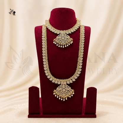 An ornate gold-toned traditional necklace displayed on a deep red velvet jewelry bust against a soft cream draped backdrop. The necklace features intricate filigree detailing, beadwork, and dangling pearl-like accents.