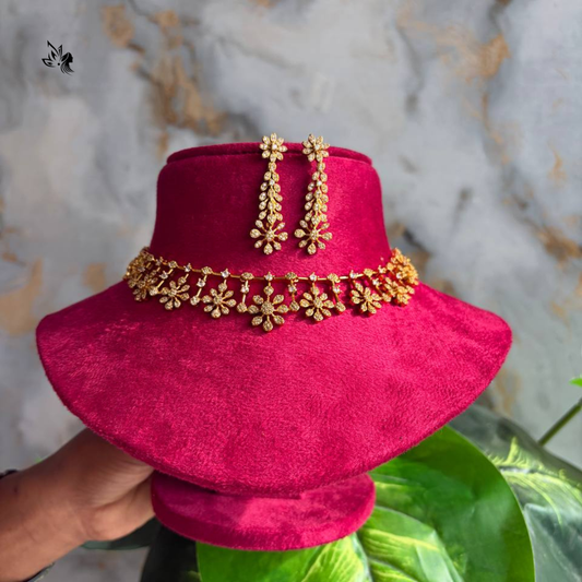 A pair of gold floral drop earrings displayed on a bright pink velvet jewelry stand, with a matching gold chain necklace draped around the neck of the stand. A hand is holding the stand on the left, and a marble-patterned wall and green leaves are in the background.