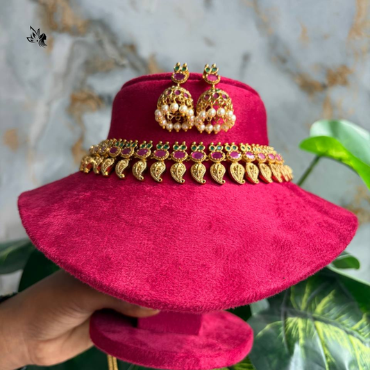 A bright pink velvet bridal headpiece with a wide brim, decorated with a gold leaf and red stone band, and styled with matching gold and pearl earrings. A hand holds the stand against a marble and green plant background.
