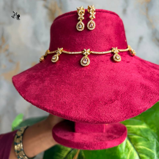 Gold teardrop pendant earrings and matching chain jewelry displayed on a magenta velvet hat stand, with a hand holding the stand in front of a marble-patterned background and green leaves.
