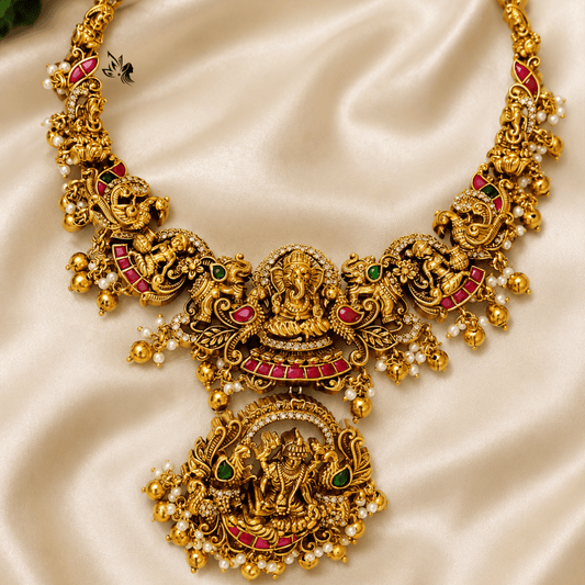 An ornate gold-toned traditional necklace with intricate filigree, red enamel accents, green stone details, and small pearl-like beads, displayed on draped cream satin fabric.